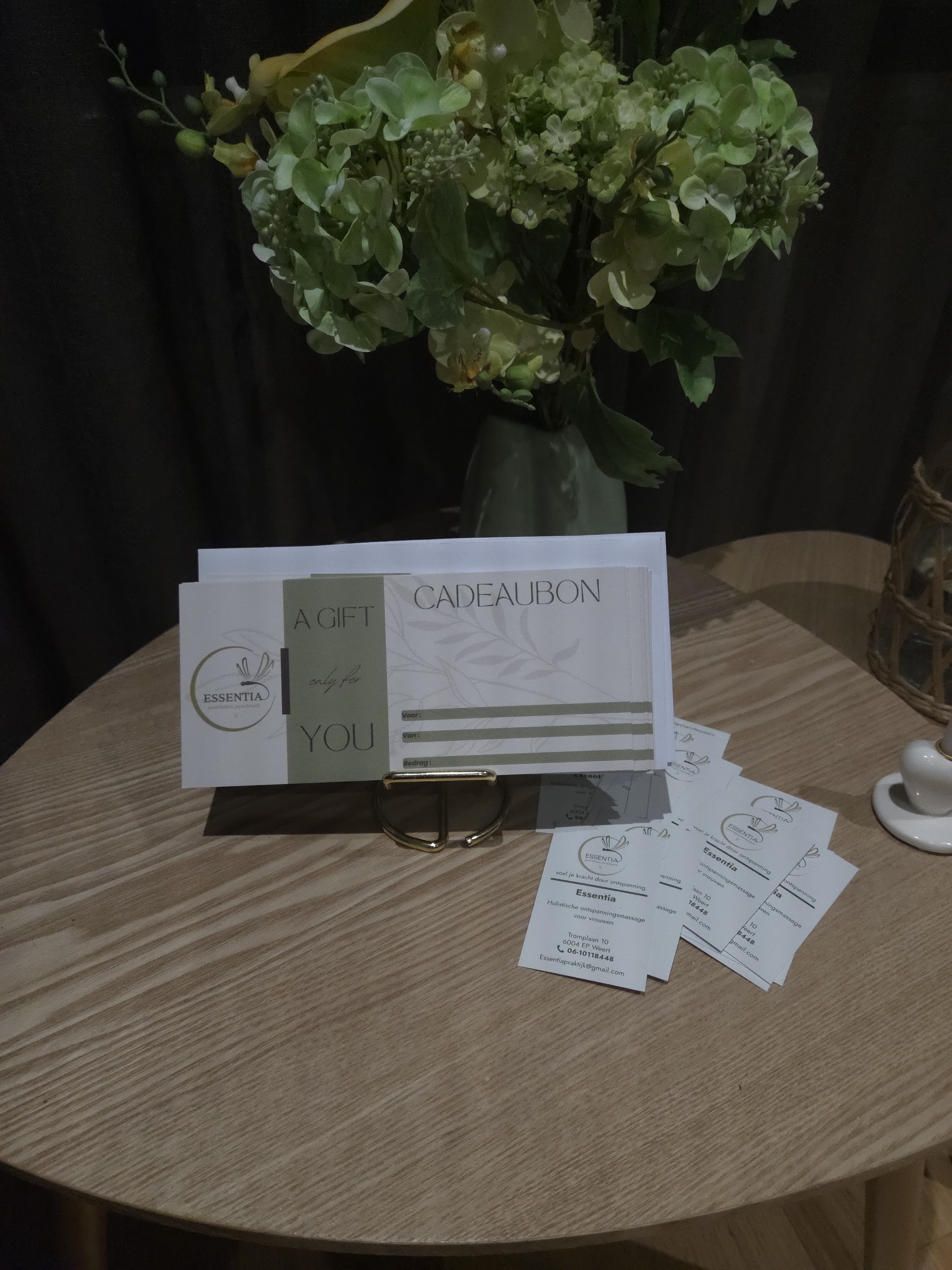 Een cadeaubon van Essentia in een gouden standaard, met visitekaartjes en een vaas met groene bloemen