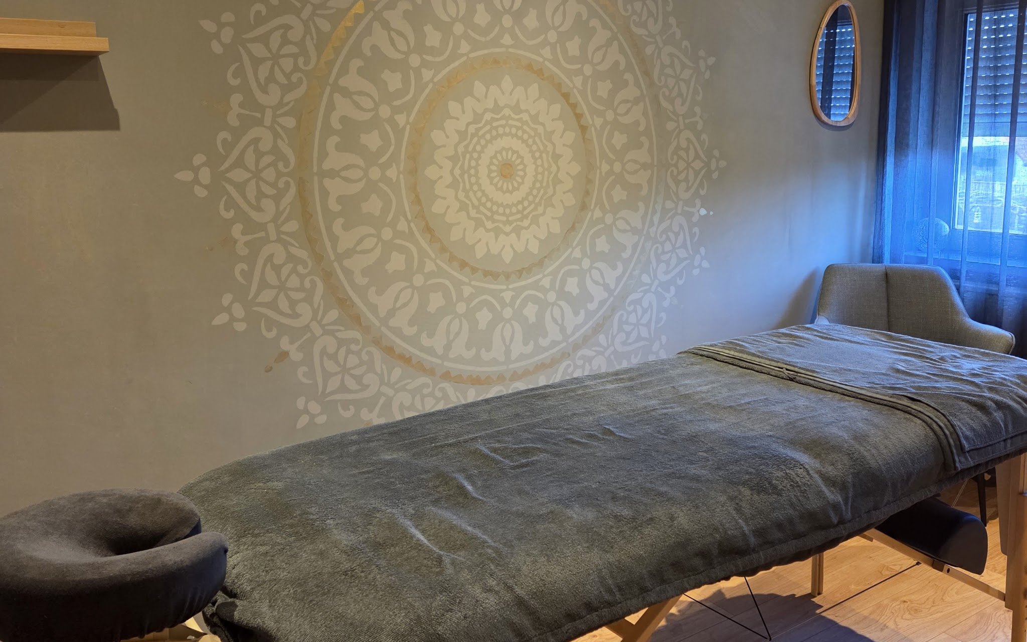 De behandelkamer van Essentia: een massagetafel met warme grijze doeken voor een muur met een wit met gouden mandala
